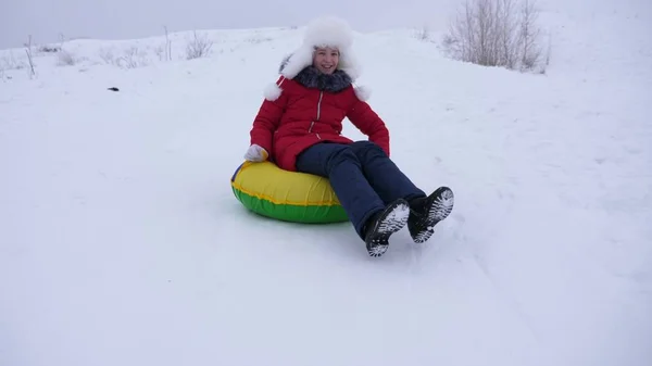 Счастливые детские слайды на снежных горках на санях, трубах. Девушка играет зимой в парке. веселая девушка-подросток в белой шляпе и красной куртке смеется — стоковое фото