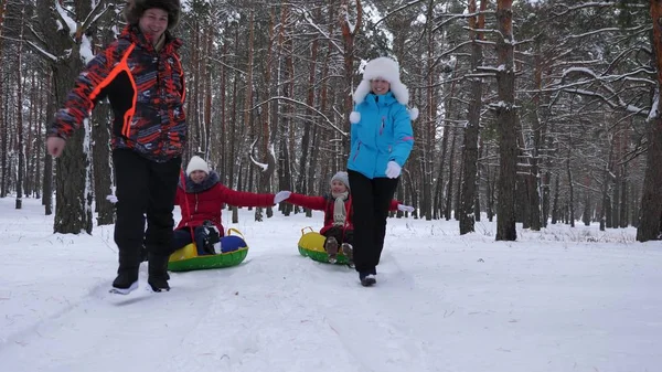Glückliche Väter und Mütter ziehen mit Kindern Schlitten im Winterwald. Eltern spielen im Winter mit Kindern in einem verschneiten Park. glückliches Familienrodeln während der Weihnachtsfeiertage. — Stockfoto