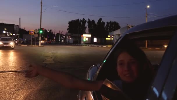 Junges schönes Mädchen winkt aus dem Autofenster. Mädchen fährt nachts im Auto durch die Stadt. Zeitlupe. — Stockvideo