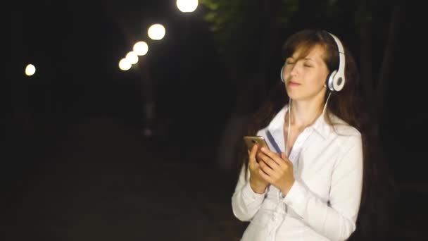 Menina em fones de ouvido e smartphone está ouvindo música, dançando e sorrindo à noite em um parque da cidade . — Vídeo de Stock