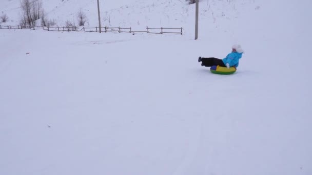 Ein fröhliches junges Mädchen rutscht im Schnee auf einem aufblasbaren Schneeröhrchen eine Rutsche hinunter und winkt mit der Hand. Mädchen spielen im Park für die Weihnachtsfeiertage im Winter. Zeitlupe. — Stockvideo