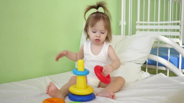 Little baby is sitting on bed in hospital ward and playing with pyramid. Sick girl is recovering in hospital setting under supervision of doctors — Stock Video