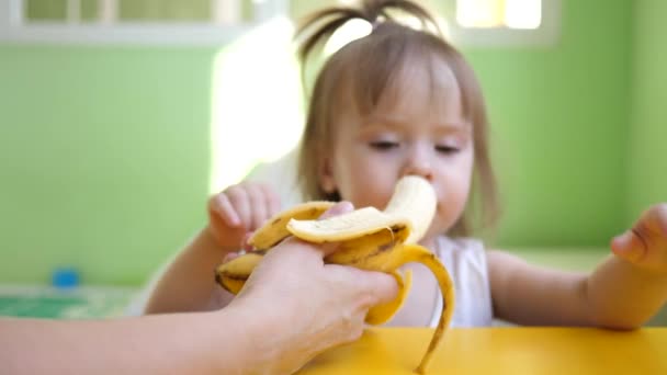 Мама кормит малыша бананом, сидя за столом на кухне. Дополнение для маленьких детей. Сладкий малыш — стоковое видео
