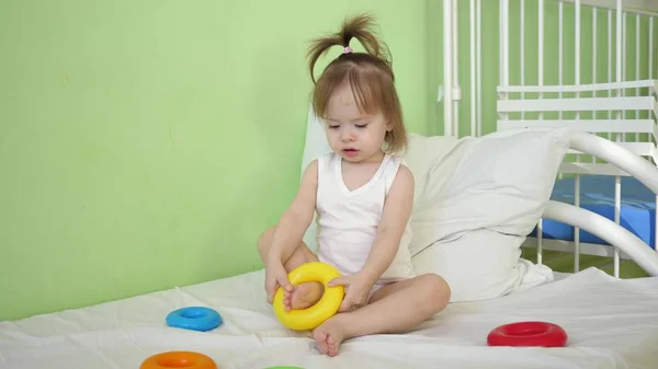 Criança coloca um anel de brinquedo em seu pé. Bebê brincando no hospital na cama com pirâmide e anéis coloridos. Brinquedos educativos para crianças pré-escolares e do jardim de infância . — Fotografia de Stock