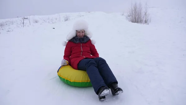 Ευτυχισμένο παιδί διαφάνειες στο χιόνι διαφανειών σχετικά με έλκηθρο, σωλήνωση. κορίτσι παίζει τον χειμώνα στο πάρκο. χαρούμενα έφηβος κορίτσι σε λευκό καπέλο και κόκκινο σακάκι έχοντας ένα γέλιο — Φωτογραφία Αρχείου