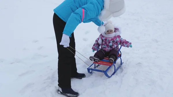 Giovane madre rotola piccolo bambino sulla slitta lungo la strada innevata in inverno. Bambino è cattivo e piange mentre seduto in slitta — Foto Stock