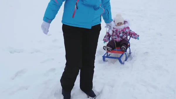 Młoda matka bułki małe dziecko na sanki wzdłuż zaśnieżonej drodze w zimie. Dziecko jest niegrzeczne i płacze siedząc w sanki — Zdjęcie stockowe
