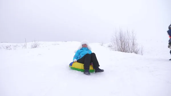 Glückliches Mädchen gleitet auf Schlitten durch den Schnee, schöne junge Frau gleitet auf einem aufblasbaren Schneeröhrchen im Schnee hinunter und winkt mit der Hand. Mädchen spielt im Park für die Weihnachtsfeiertage im Winter — Stockfoto