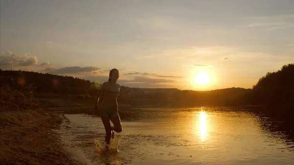 Giovane ragazza che corre lungo la spiaggia spruzzando acqua da sotto i piedi, bel tramonto sul fiume, riprese al rallentatore — Foto Stock