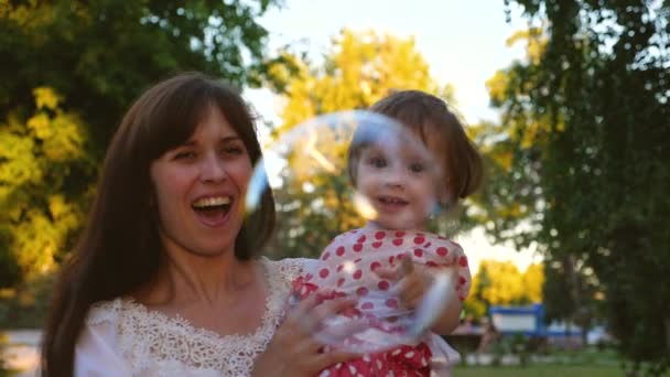 Glückliche Mutter und Tochter fangen große Seifenblasen im heißen Sommerpark und lachen. Kindermädchen mit kleinem Mädchen spielt spazieren und lächelt — Stockvideo