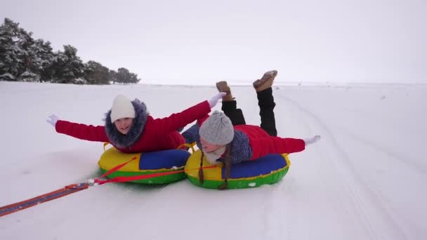 Glada barn tonåringar rulla på snöiga plattan längs vit snöig väg och skratta. Glada flickor gå kälkåkning på vintern och leende. Jullov — Stockvideo