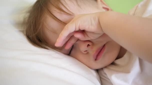 Encantador bebé se duerme en la cama blanca en su cama en la habitación en casa. concepto de niño dormido. niño quiere dormir y se frota los ojos con las manos. El niño duerme en la sala del hospital. primer plano — Vídeo de stock