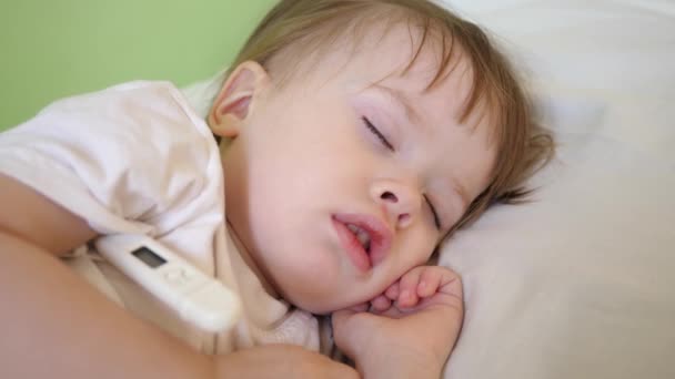 Μικρό παιδί κοιμάται στο νοσοκομείο ward λευκά κλινοσκεπάσματα και μετρά τη θερμοκρασία με θερμόμετρο. Θεραπεία των παιδιών στο νοσοκομείο. Άρρωστο βρέφος βελτιώνει την υγεία του στο νοσοκομείο — Αρχείο Βίντεο