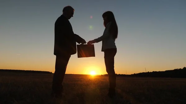 Силуэт бизнесмен и молодая деловая женщина заключили сделку, передают друг другу черный портфель и пожимают друг другу руки на фоне заката на голубом небе — стоковое фото