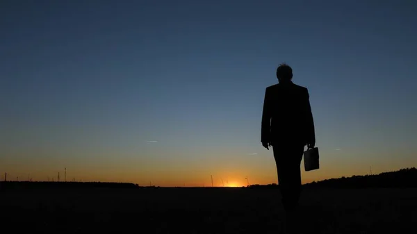 Silhouette di uomo d'affari con valigetta che cammina attraverso il campo al tramonto, contro un cielo blu — Foto Stock