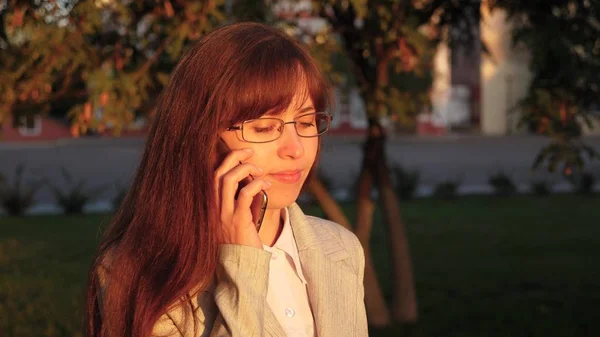 Mujer de negocios con gafas en traje de luz va a trabajar. mujer camina por una calle de la ciudad iluminada por rayos de sol y habla en un teléfono inteligente . — Foto de Stock