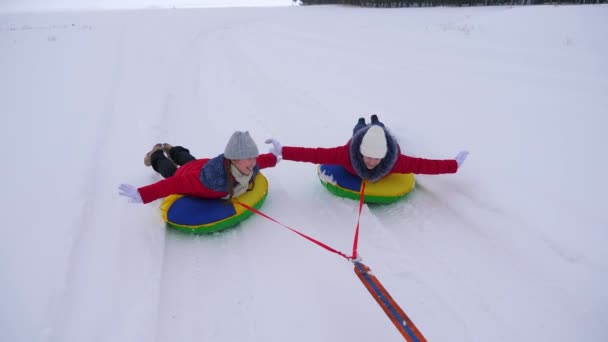 Teen girls roll on sleigh ride along snow white road in winter. Girlfriends ride snow saucer in snow. Christmas Holidays — Stock Video