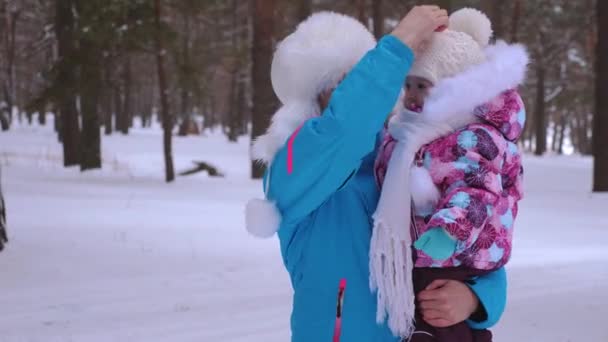 Η μαμά κρατάει τη μικρή της κόρη στην αγκαλιά της και ισιώνει το χειμώνα πάρκο πεύκων καπέλο. Υγιή περίπατοι για ψυχρός ημέρα υγείας μεταξύ γονέα και μωρού. Μαμά και το παιδί περπατά μέσω κωνοφόρων χιονισμένο δάσος — Αρχείο Βίντεο