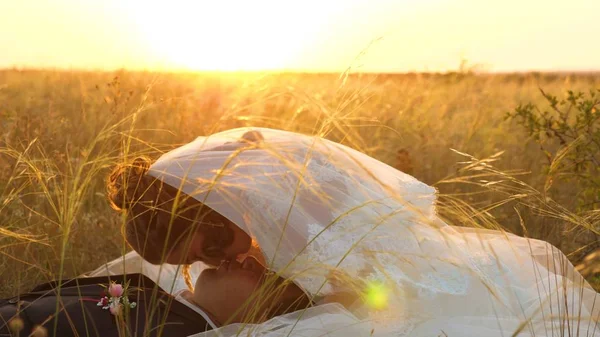 Sposa accarezza sposo con erba stelo in faccia. famiglia felice è sdraiato in campo in raggi di bel tramonto. giovane donna bacia il suo amato uomo sulle labbra la sera d'estate sull'erba. famiglia felice — Foto Stock