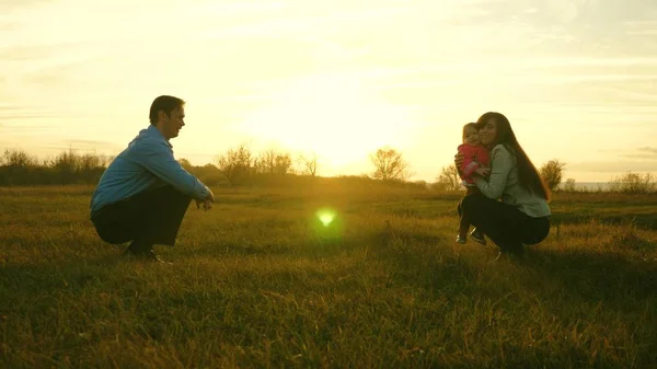 Baby gaat op gazon van vader naar moeder. kind neemt eerste stappen in het park. Mama en papa spelen met kind op het gras bij zonsondergang. familie geluk concept — Stockfoto