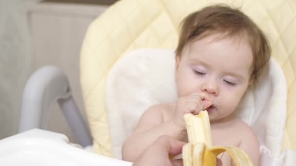Kleines Baby isst eine Banane im Stuhl in der Küche. — Stockvideo