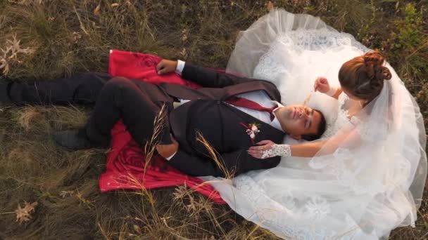 Sposa e sposo al tramonto riposano insieme seduti nel campo con il grano e si sorridono a vicenda. Luna di miele. Amanti uomo e donna. Relazioni coniugali — Video Stock