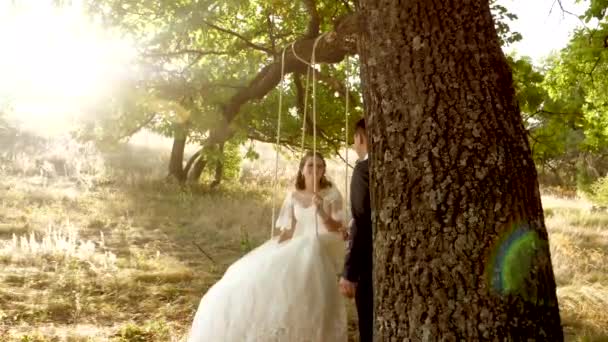 Groom secoue sa mariée sur swing et sourit avec un regard aimant. Femme et homme en réunion romantique sont heureux dans les moments forts du coucher du soleil — Video