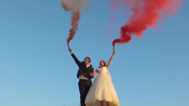 Lyckliga bruden och brudgummen viftande färgade rosa rök mot blå himmel och skrattar. Smekmånad. Romantik. Förhållandet mellan man och kvinna — Stockvideo