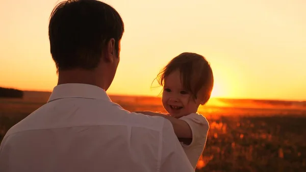 Buon papà e figlioletta seduti tra le sue braccia al tramonto sole splendente. La ragazza ride giocando con papà durante la passeggiata serale. Primo piano — Foto Stock