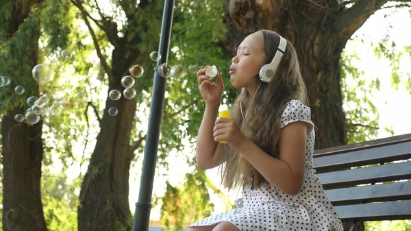 Dospívající dívka v sluchátka sedí na lavičce a fouká mýdlové bubliny a úsměvy. — Stock fotografie