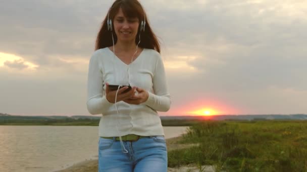 Chica feliz viendo vídeo en la tableta y caminando en el parque junto al lago. hermosa chica en los auriculares camina en la playa con la tableta y es la videoconferencia en línea . — Vídeos de Stock
