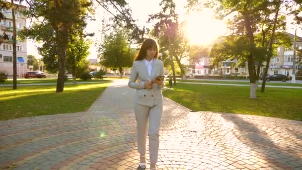 Hermosa mujer de negocios camina por el callejón en el parque con la tableta en sus manos, chica en un traje de negocios ligero va a trabajar . — Vídeo de stock