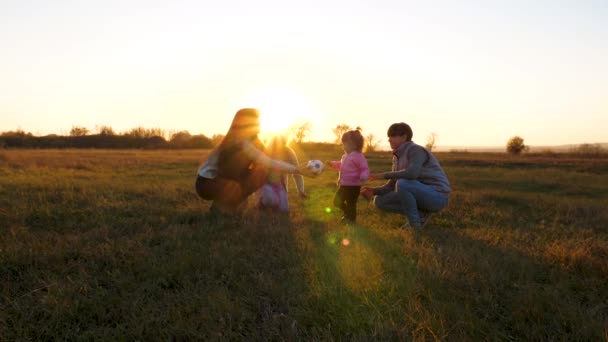 Mamá juega con su pequeña hija bola de bebé. niño pequeño jugando pelota con los niños y mamá. Familia jugando con un niño pequeño en una pelota de fútbol infantil en el parque al atardecer. Movimiento lento — Vídeos de Stock