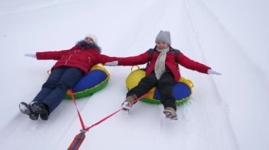 Happy teen girls go but snowy road in winter snowy mountain, straightening their arms in flight and laughing. Happy childhood in winter on sled. Christmas Holidays