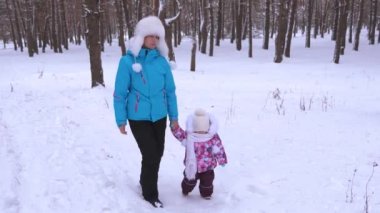 Nanny leads hand of little girl on snowy road while walking in fresh air in park in winter. Mom and daughter make first steps in winter forest