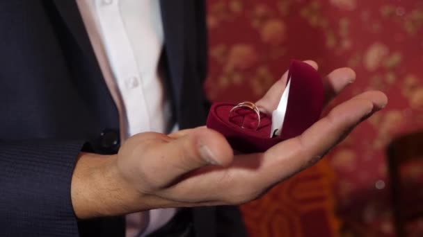 Novio sostiene en su caja de mano de anillos de bodas de oro. hombre con un traje con anillos de oro en la mano. primer plano . — Vídeos de Stock