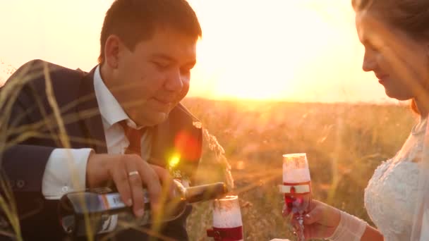 Noivo derrama deliciosos copos de vinho no campo em raios brilhantes de sol para a noiva. close-up. os recém-casados felizes bebem a champanha no prado em raios de um belo pôr-do-sol. conceito de casamento feliz — Vídeo de Stock