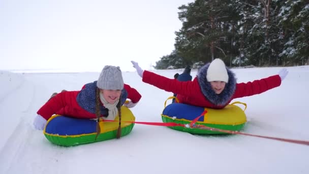 Happy children have fun riding snow saucer and laugh on snowy winter road on winter frosty day. Teens play on sled in winter field and smile. Games in fresh frosty air. Christmas Holidays — Stock video