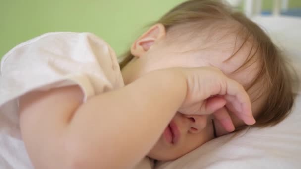 Όμορφο μωρό πέφτει να κοιμηθεί σε λευκό κρεβάτι σε κρεβάτι του στο δωμάτιο στο σπίτι. έννοια του ύπνου του παιδιού. θέλει να κοιμηθεί και να τρίβει τα μάτια του με τα χέρια του παιδιού. το παιδί κοιμάται στον θάλαμο νοσοκομείου. γκρο πλαν — Αρχείο Βίντεο