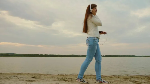 Ragazza in cuffia cammina in serata sulla spiaggia con un tablet e ascoltare musica . — Foto Stock