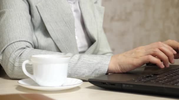 Chica sentada en silla en la oficina y escribiendo en el ordenador portátil. taza de café blanco está en el escritorio de la oficina. joven mujer de negocios que trabaja en la computadora. primer plano — Vídeos de Stock