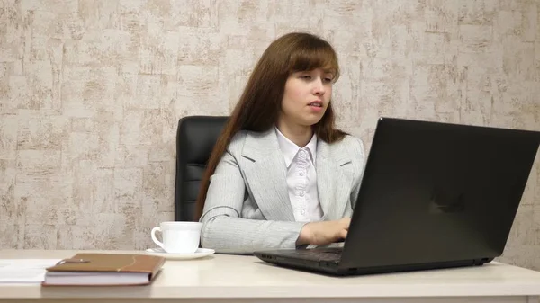 Piękna dziewczyna siedzi w fotelu w biurze i pisania na laptopa i picia kawy z białego kubka. Młoda kobieta kobieta interesu pracę przy komputerze. dziewczyna jest na czacie na laptopie — Zdjęcie stockowe