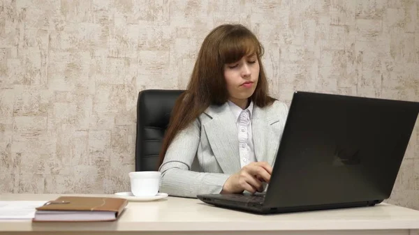 Красива дівчина сидить у кріслі в офісі і друкує на ноутбуці і п'є каву з білої чашки. молода жінка бізнес-леді працює за комп'ютером. дівчина спілкується на ноутбуці — стокове фото