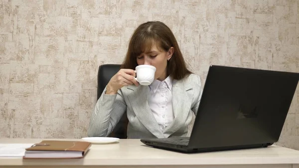 Vacker flicka sitter i stol på kontoret och skriva på laptop och dricka kaffe ur vit kopp. ung kvinna affärskvinna arbetar vid dator. flicka chattar på en bärbar dator — Stockfoto