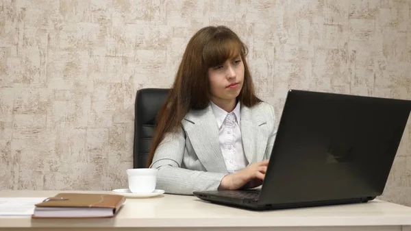 Menina bonita sentada na cadeira no escritório e digitando no laptop e beber café de copo branco. mulher jovem empresária que trabalha no computador. menina está conversando em um laptop — Fotografia de Stock
