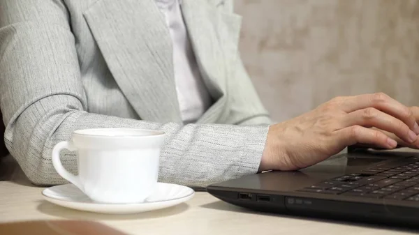 Flickan sitter i stol på kontoret och skriver på laptop. vit kaffekopp står på kontoret skrivbordet. ung kvinna affärskvinna arbetar vid dator. närbild — Stockfoto