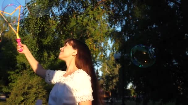 Chica sopla grandes burbujas de jabón y se ríe mientras camina en el parque. En cámara lenta. Paseos de verano y juegos en la naturaleza . — Vídeos de Stock