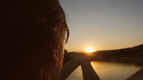 Молодая девушка тянет руки к солнцу, закат на фоне реки — стоковое видео