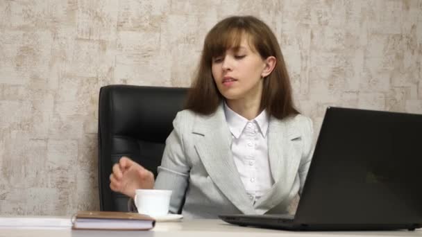 Menina bonita está sentada na cadeira no escritório e beber café, chá, e está conversando em um laptop. mulher jovem empresária trabalha no computador . — Vídeo de Stock