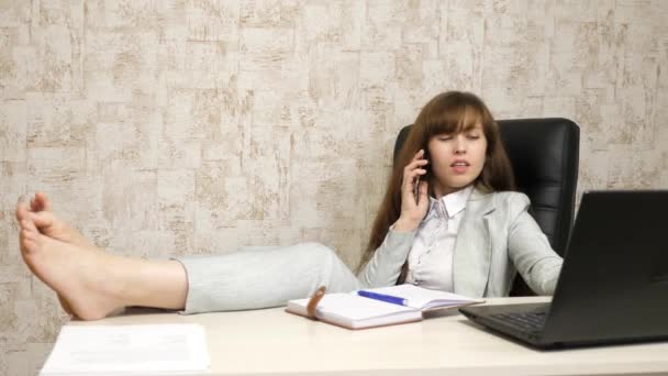 Bela mulher de negócios sentada na cadeira com os pés descalços na mesa. menina no trabalho no escritório no telefone à mesa e descansando . — Vídeo de Stock
